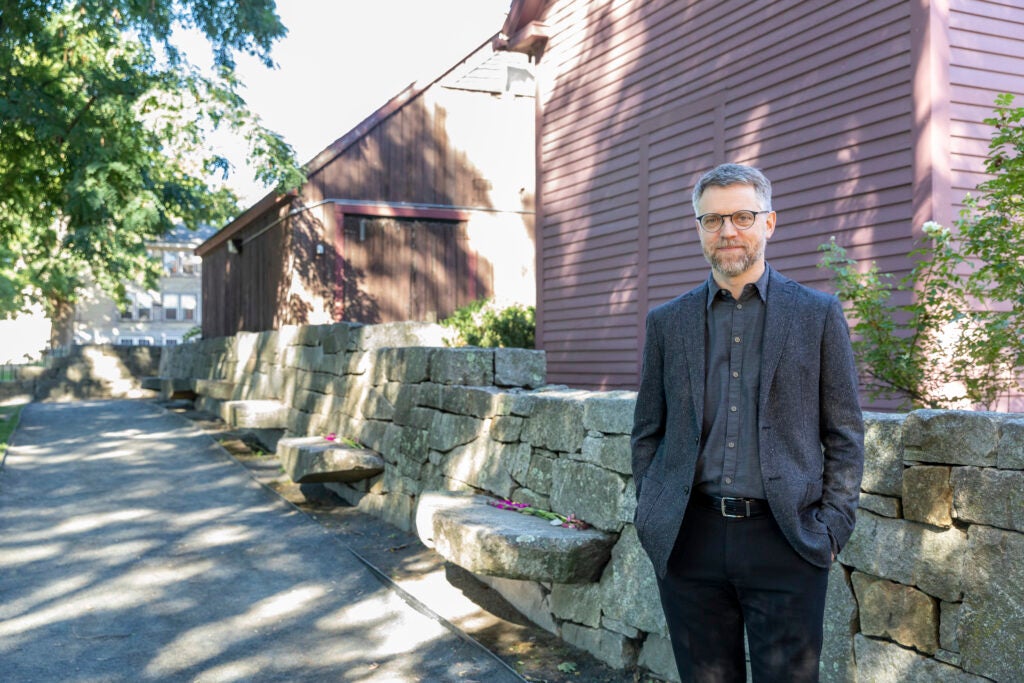 Dan Lipcan, Director of PEM’s Phillips Library, stands in front of the Salem Witch Trials Memorial in Salem, Massachussetts. He is wearing a dark outfit and is standing in front of the old, red-slatted houses. 