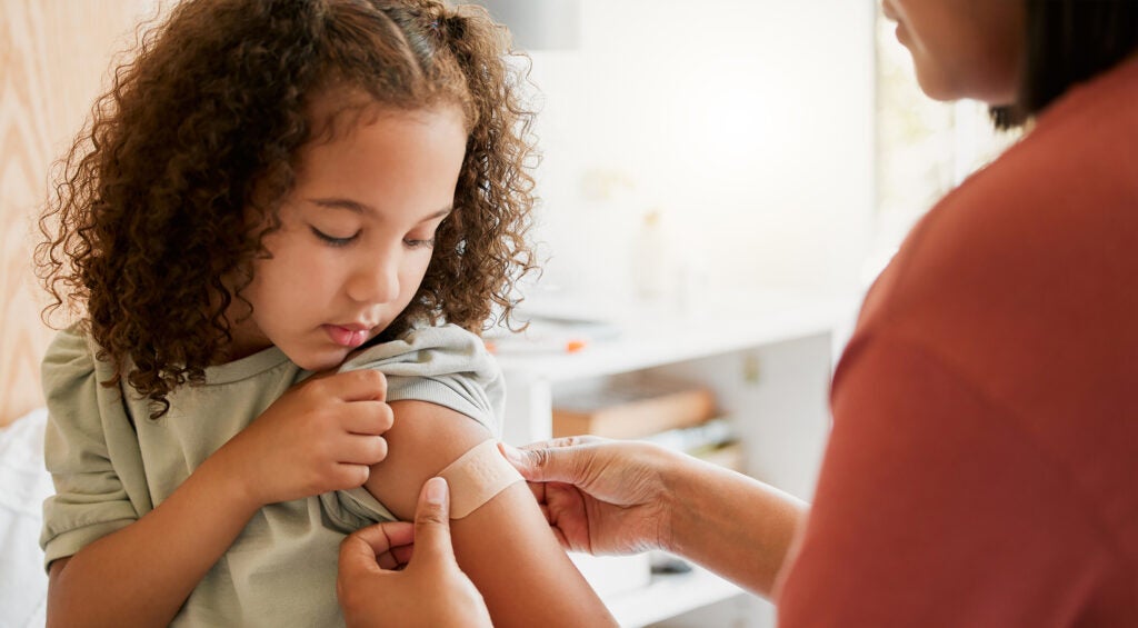Nurse putting a bandage on a child's arm after vaccinating her. Child has dark brown curly hair and wears a light green shirt. 