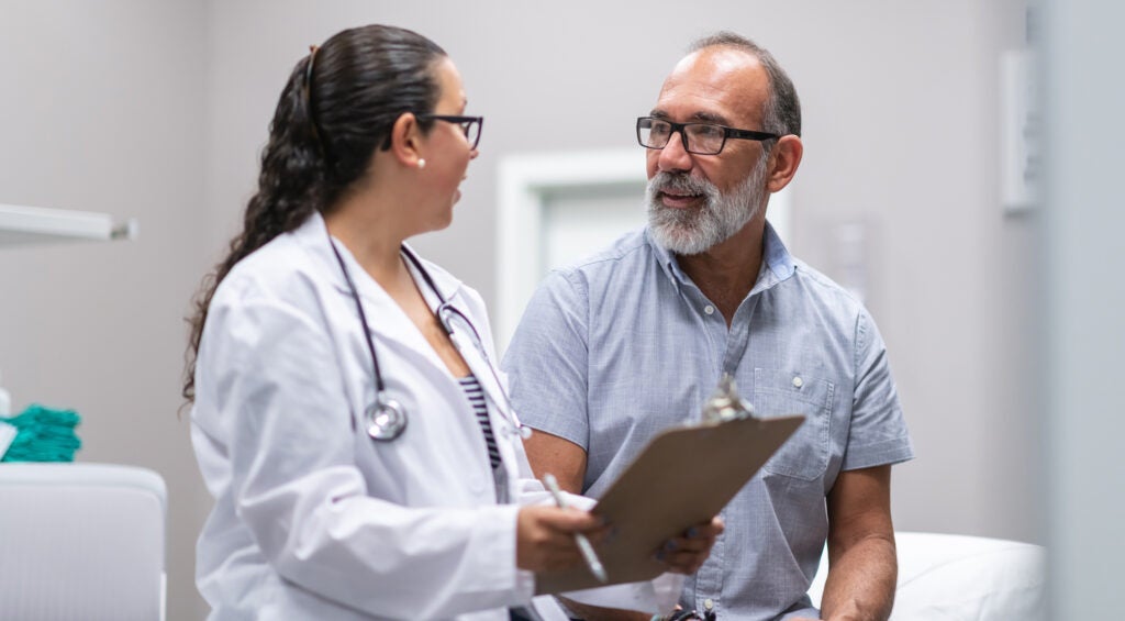 Hispanic, female doctor consults with senior male patient while they sit facing each other in a medical clinic examination room. The doctor is showing the patient test results on a clipboard.