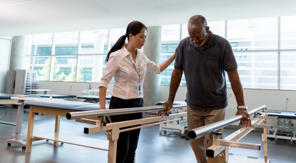 A physical therapist guides and encourages an elderly veteran as he walks using handrails. They're surrounded by physical rehabilitation equipment in a physical therapy office.