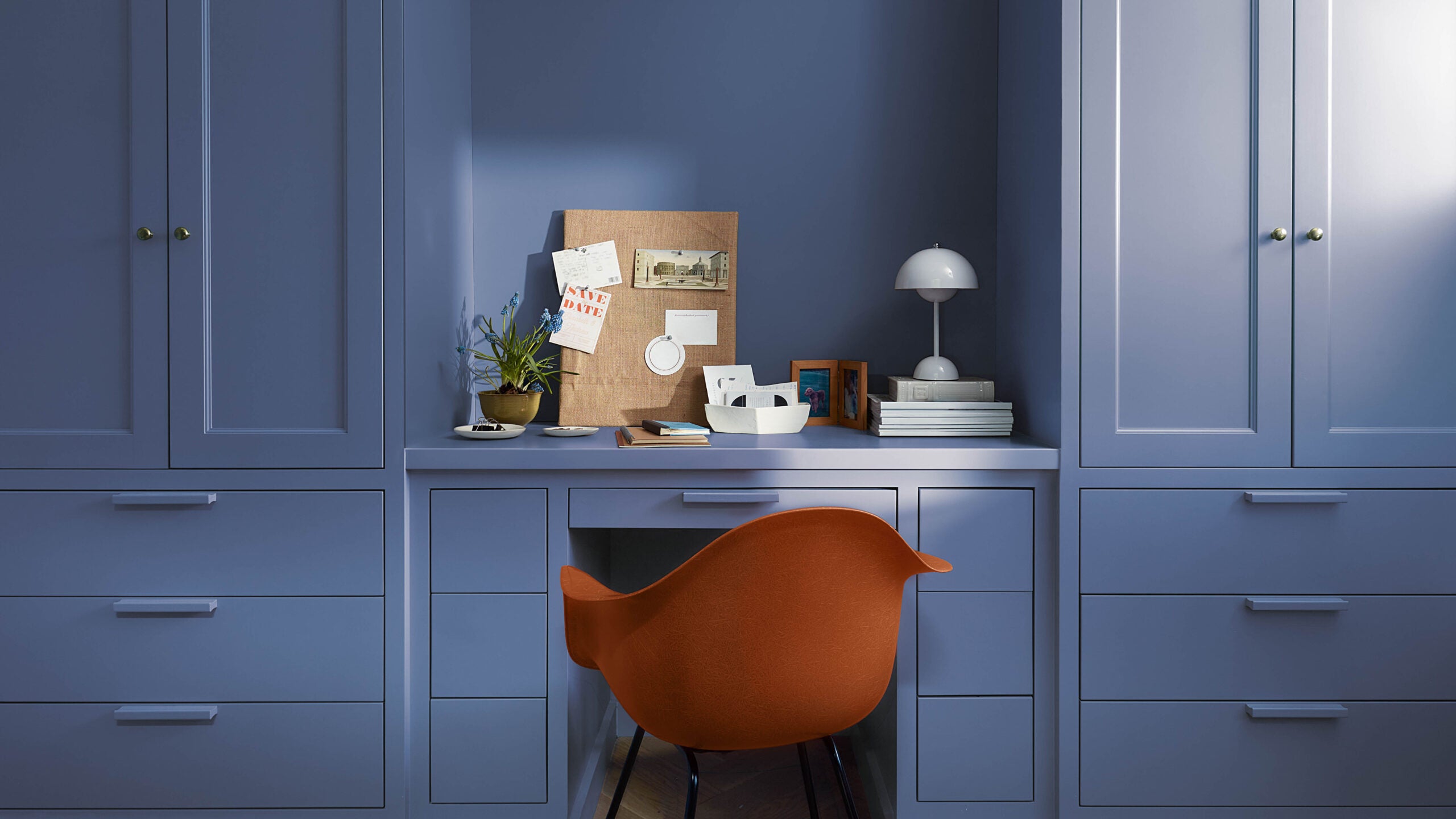 An orange chair sits at a desk built into the dark blue wall with cabinets on either side, and a few related items on the desk. 
