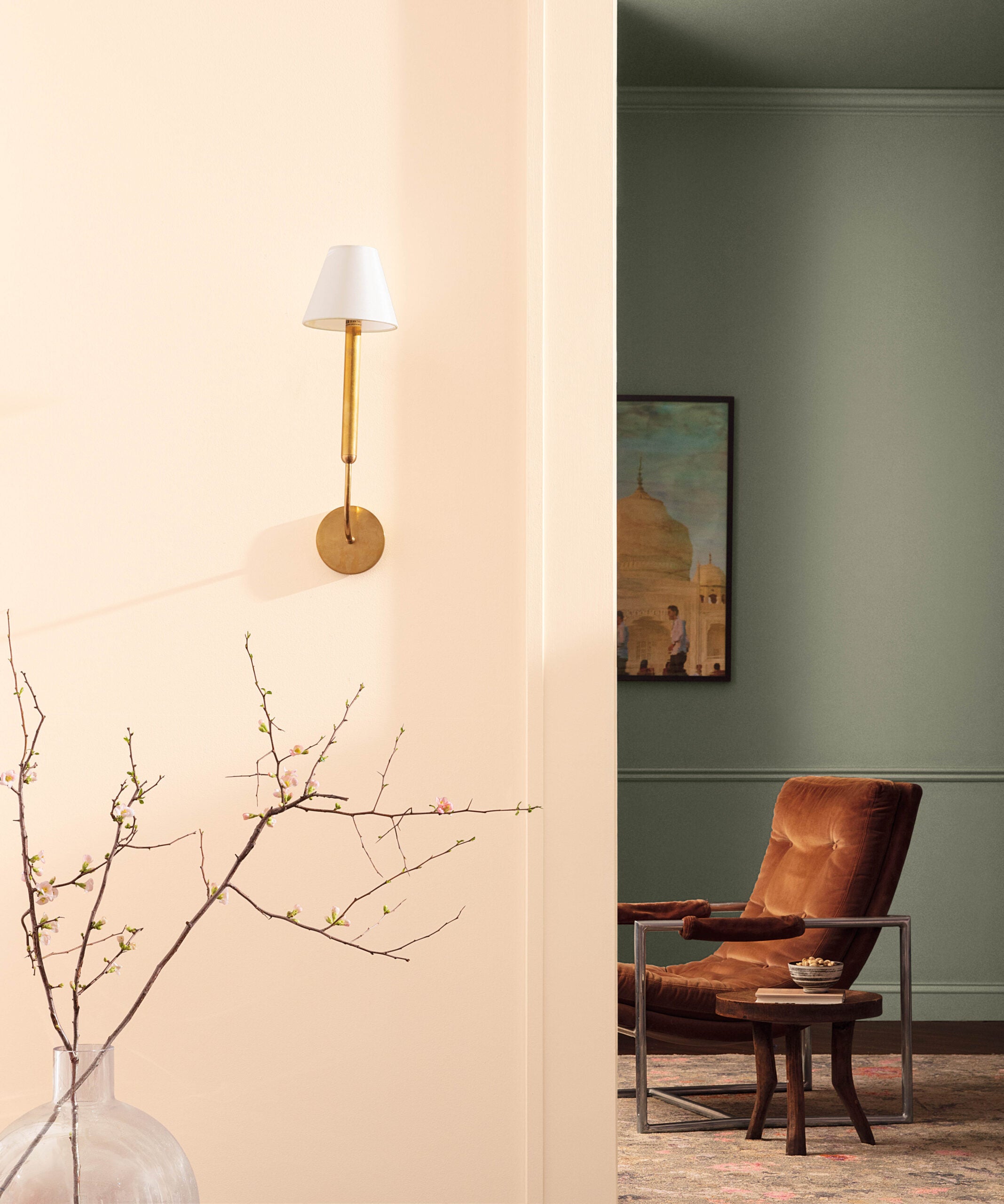 A white wall with a gold wall sconce affixed to it stands next to a background wall painted in antique pewter (a green-grey hue). Half a painting and a suede chair are visible from this angle.