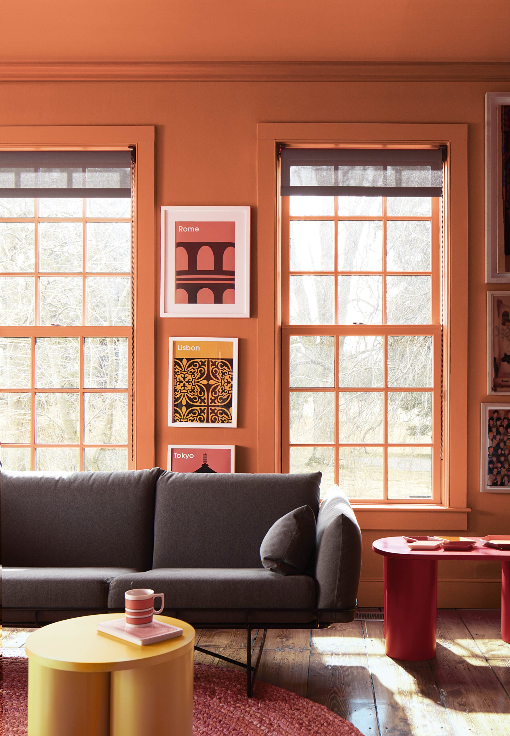 A living room with topaz orange colored walls and sun streaming through the windows contrasts against a grey couch, three paintings arranged in a line down the wall, and a few fun-colored coffee tables. 