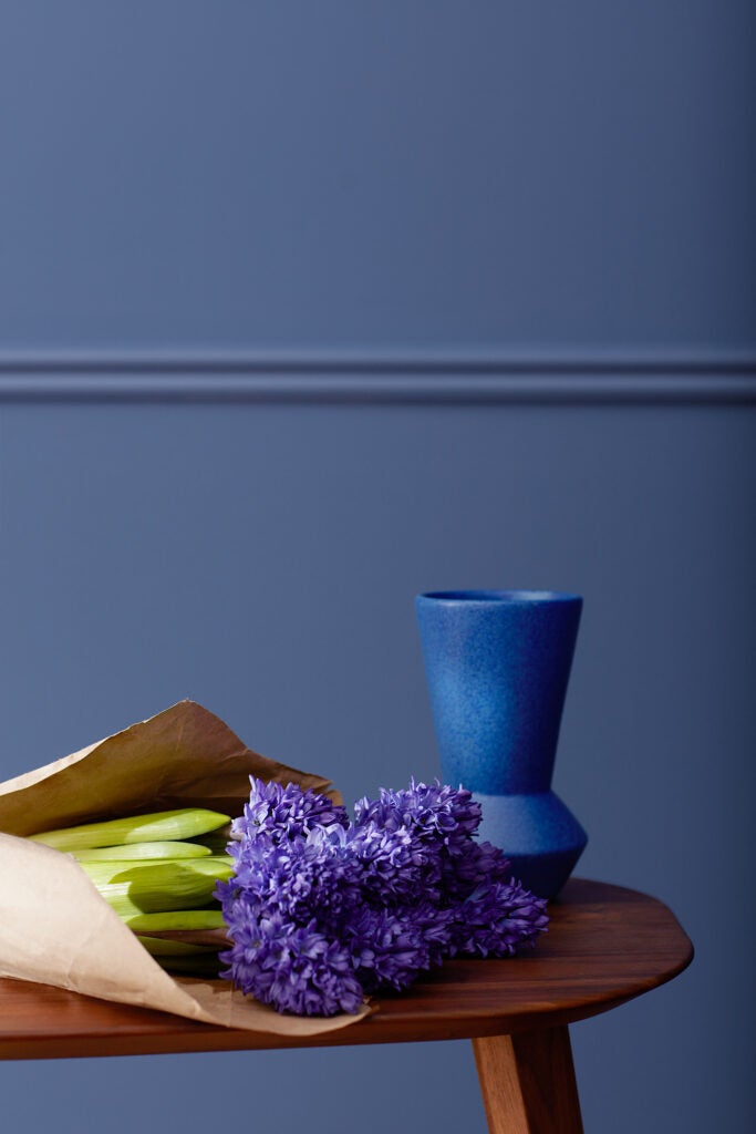 A still life of a dark blue vase and a bouquet of lilacs wrapped in brown paper supported by a wooden bench. The wall behind them is painted in blue nova. 