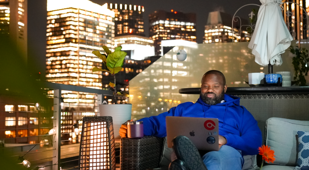 Marcus Williams sits outside at night on his balcony, looking at his computer and smiling. In the background, the Boston skyline is lit up. 