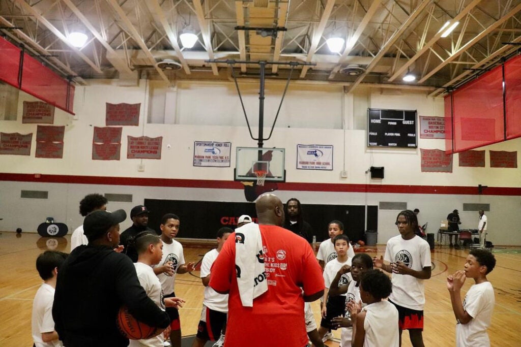 A man in a red athletic shirt coaches a team of young athletes while standing on a basketball court.