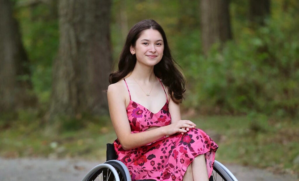 A teenage girl with long dark brown hair (Haley Carroll) wearing a sleeveless pink floral dress and a dainty necklace sits in a tennis wheelchair with her hands folded in her lap. The background is a vibrant green forest landscape.