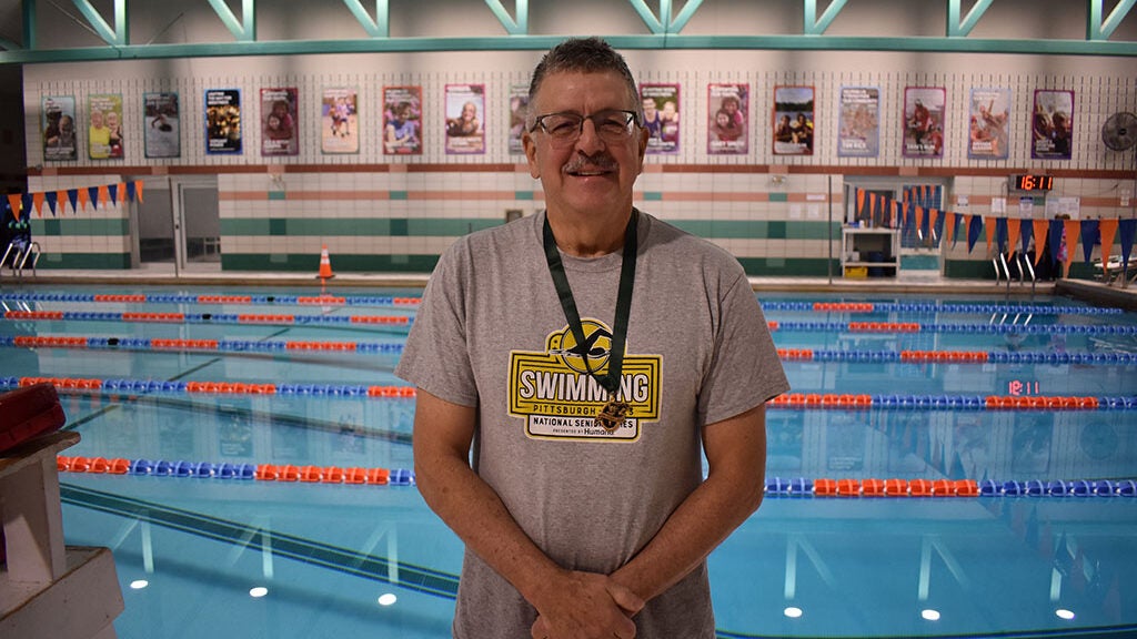 Man in a grey shirt wearing a medal standing in front of an indoor swimming pool.