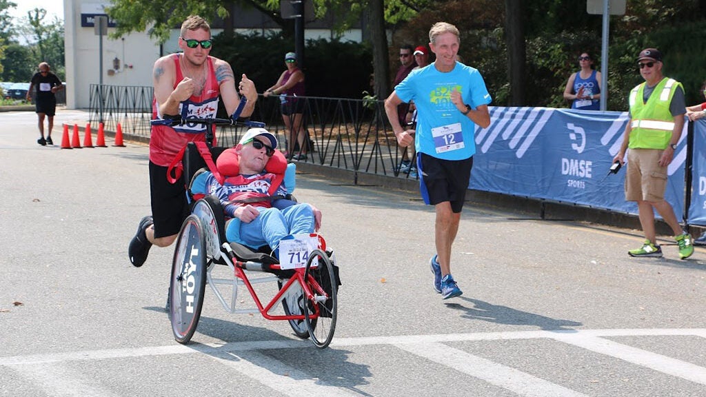 A man running and pushing the wheelchair of another man during a marathon surrounded by another runner and spectators behind a barricade. 