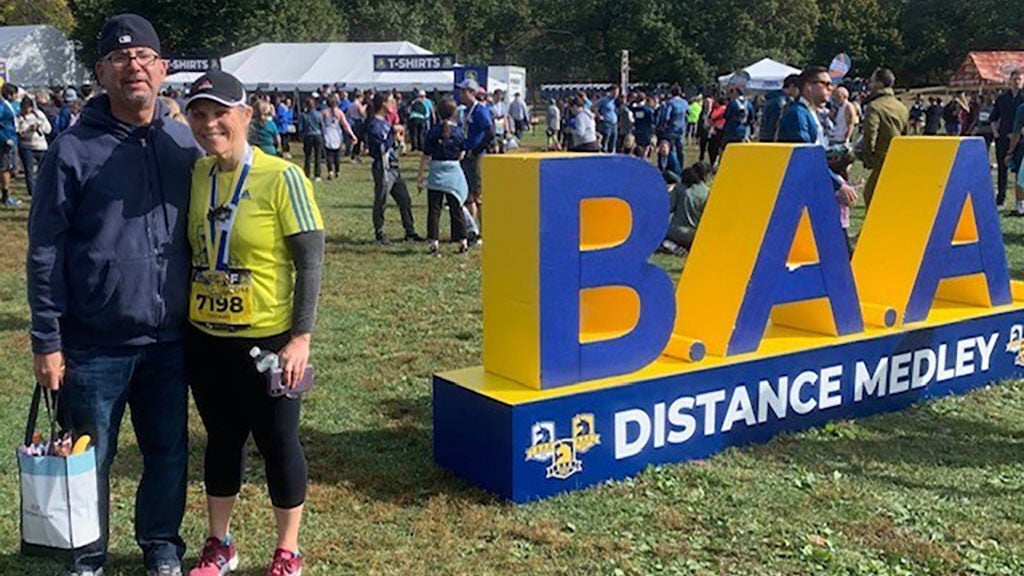 A man and a woman, wearing a race medal, smile for a photo in front of a large sign that says "B. A. A."