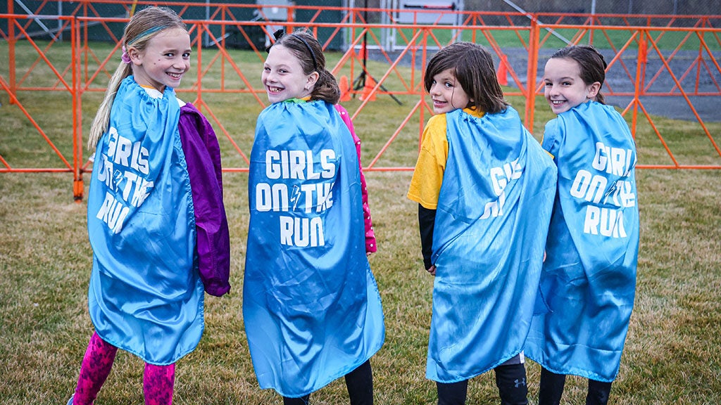 4 girls stand next to each other to smile at the camera with their backs showing off their "Girls on the Run" capes.