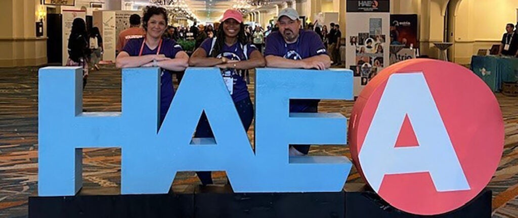 Three people stand in front of a large sign that reads the letters "H", "A", "E", "A".