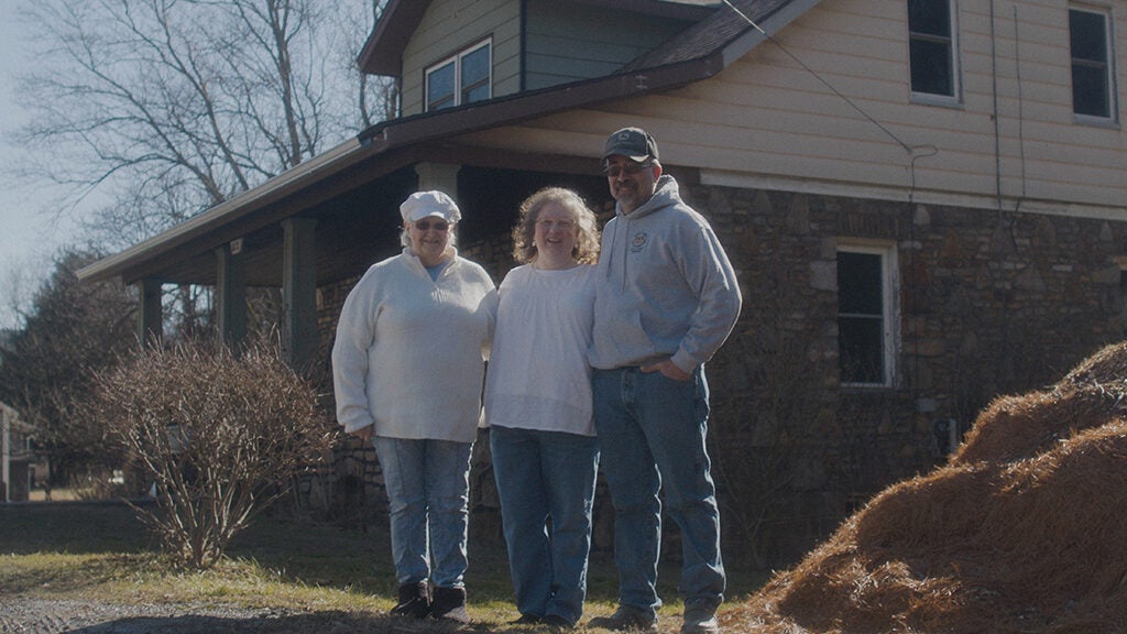 Two woman and a man stand arm and arm in front of a house, posing for a picture.