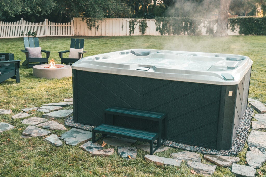 A Sundance hot tub with a dark green exterior sits atop a rock path in the middle of a grassy backyard. 