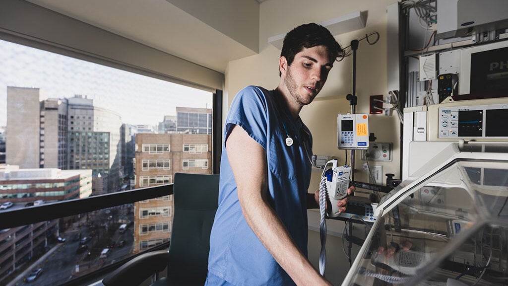 A man wearing blue scrubs stands in front of a window and checks on the NICU.