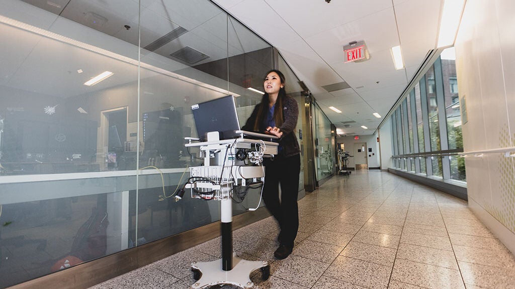 A woman wearing a dark black outfit walks down the hospital hallway pushing a machine that has a laptop on it.