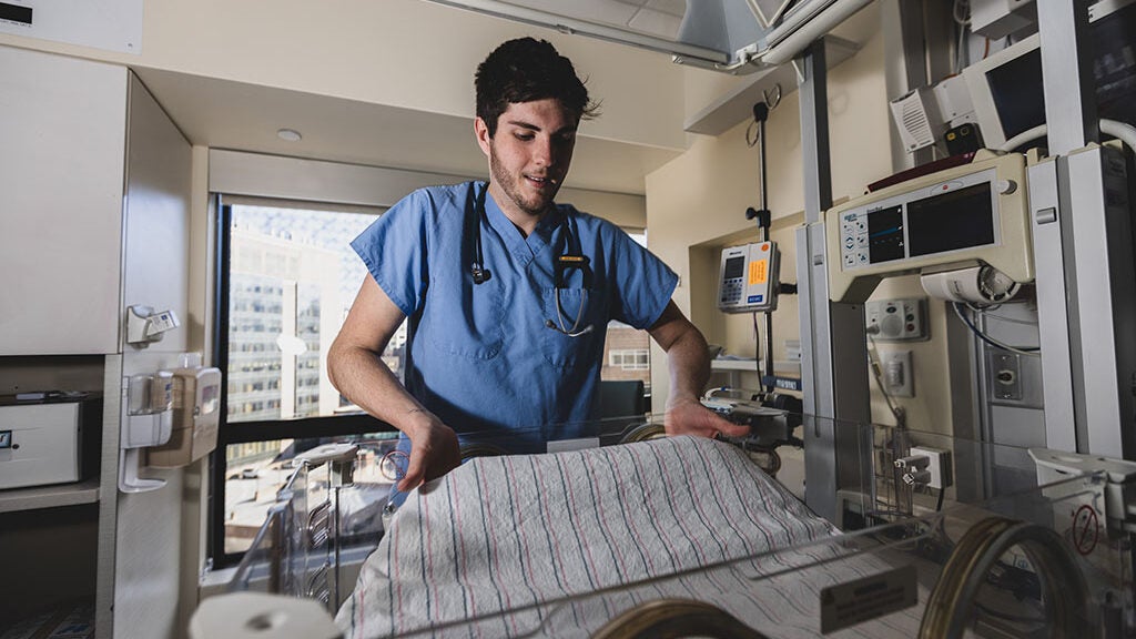 A man wearing blue scrubs adjusts the blanket in a NICU crib in a hospital.