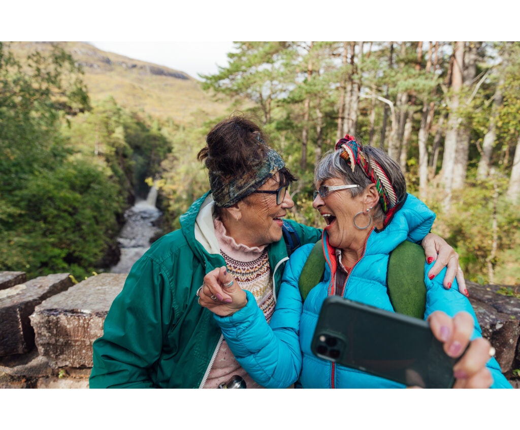 An older adult woman wraps her arm around her friend and shares a laughing, gregarious moment as they take a selfie during a hike on a scenic mountain top.