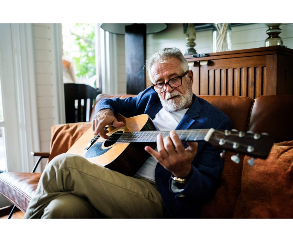An older adult man with a salt and pepper goatee, wispy white hair, and brownish-red rectangular glasses playing acoustic guitar in his living room. 