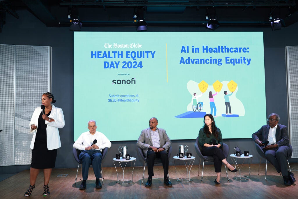 A woman wearing a white blazer and black dress addresses a crowd while four panelists sit behind her on stage. 