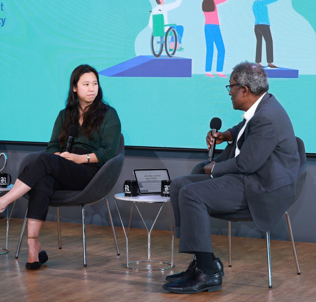 A woman wearing an emerald green blouse sits with her legs crossed on stage while a man wearing a gray suit asks her a question with a microphone. 