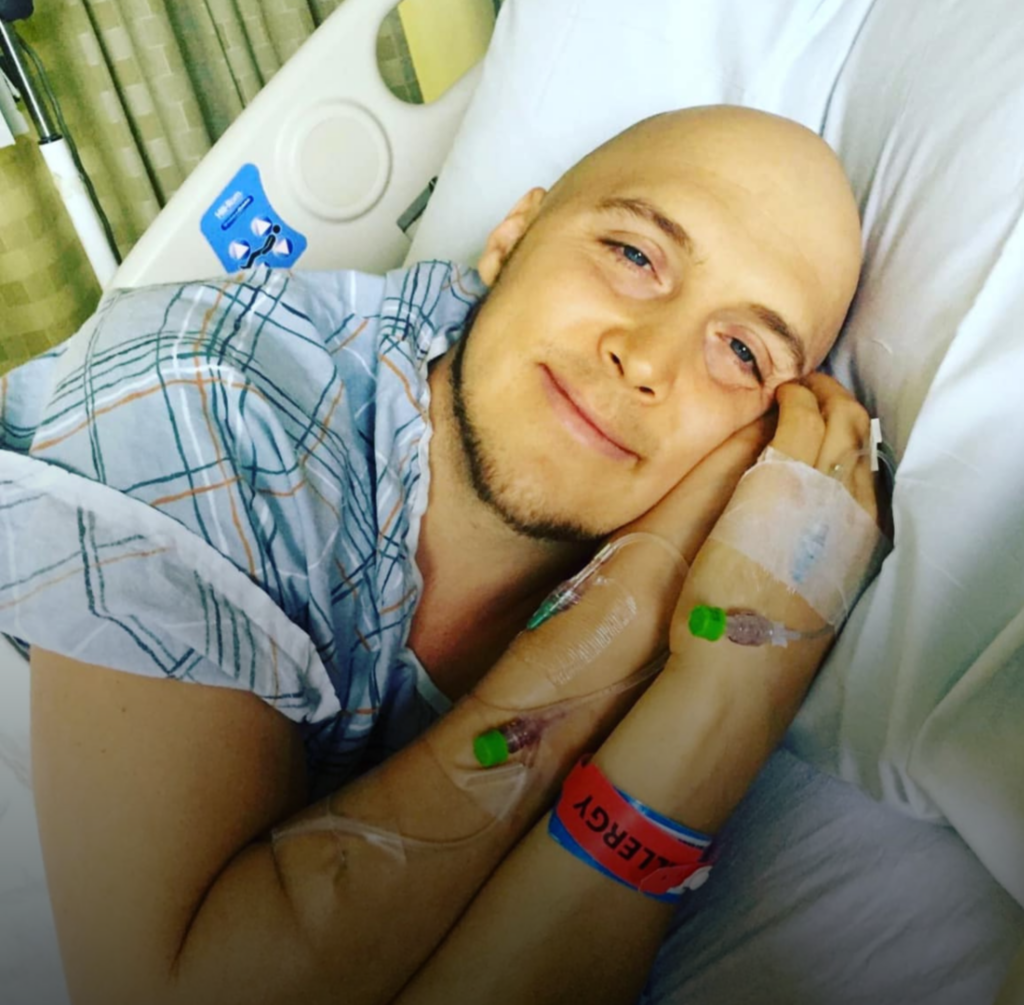 A patient with no hair in a hospital bed looking at the camera with a soft smile.