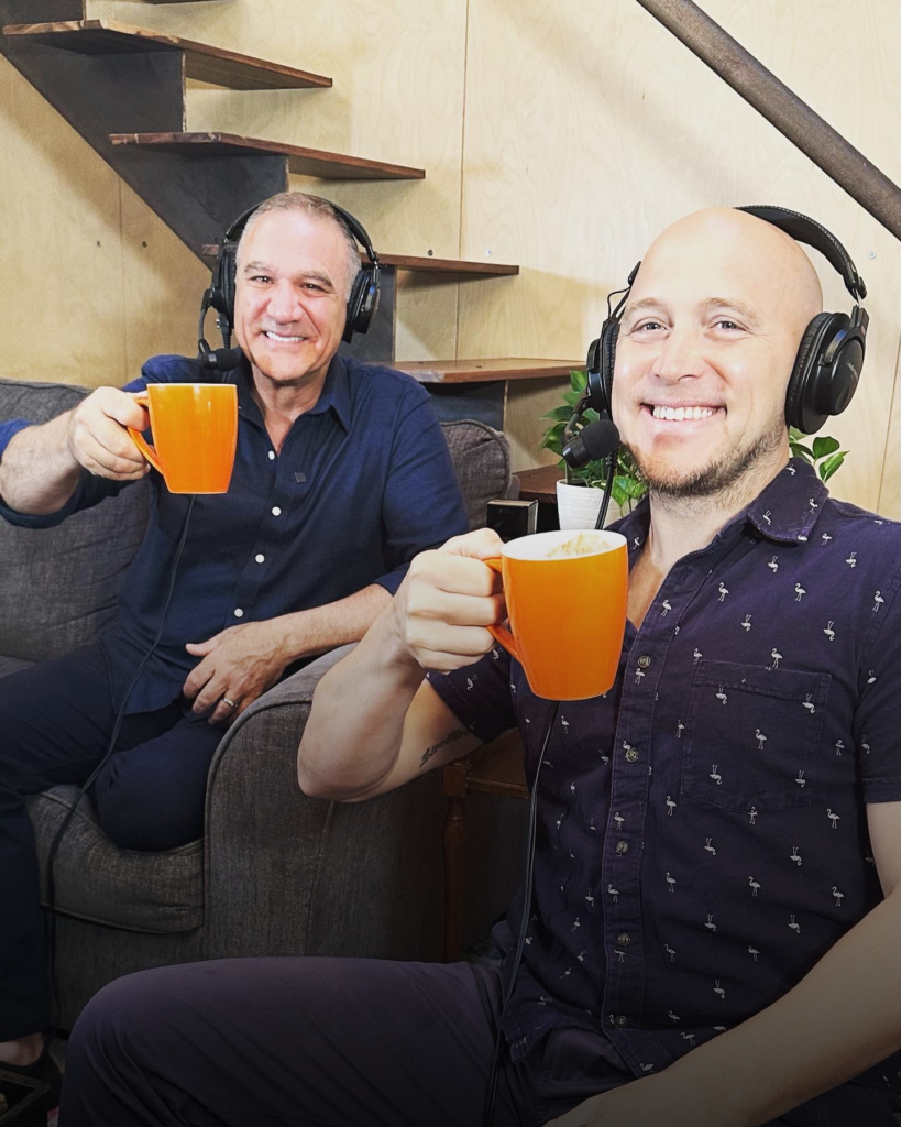 Two men wearing headsets holding up orange coffee mugs on the set of a podcast.