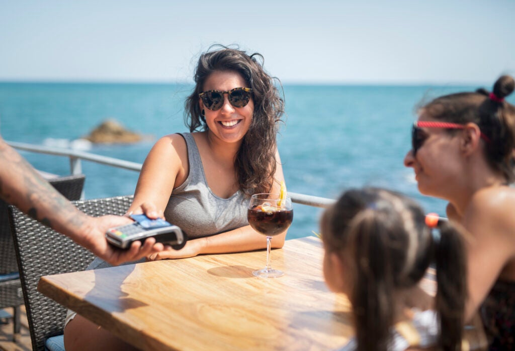 A woman pays for a meal by tapping her card at a restaurant overlooking the ocean. 