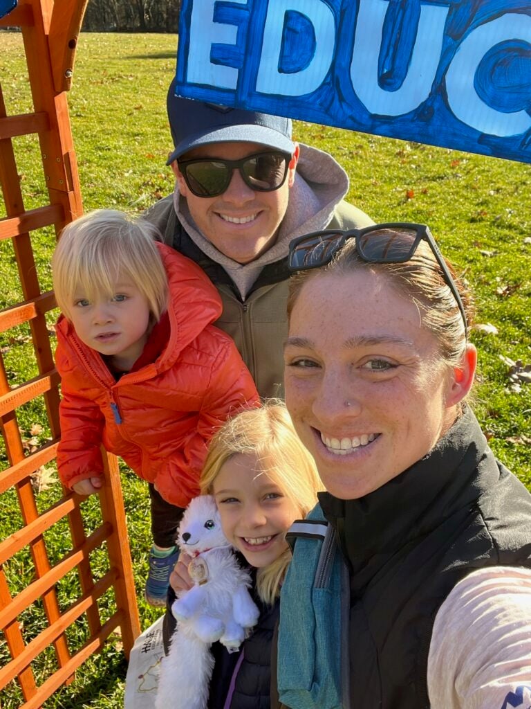 Ashley Liffers takes a selfie with her husband, smiling with their two young kids. Her daughter hugs a white dog stuffed animal. They are on a grassy field outside in the sun.