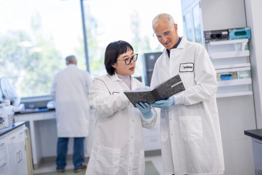 Two scientists, one male and one female, wearing white lab coats lean over a note pad while discussing in a laboratory. 