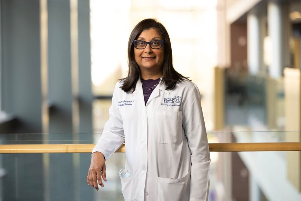 A middle-aged woman with medium-length brown hair wearing glasses and a white lab coat poses by a railing in a university building. 