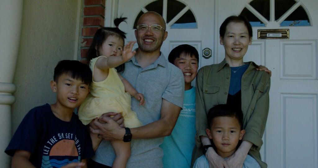 A mother and father pose smiling with their four children outside the front door to their house. 