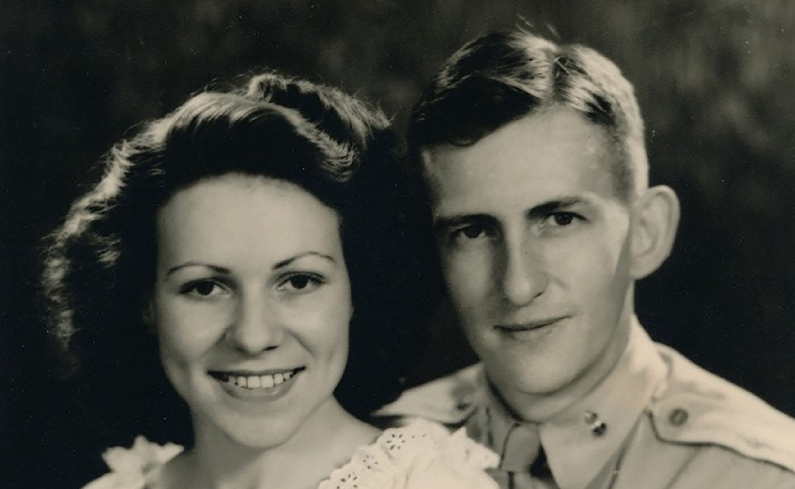 Vintage black-and-white portrait of a smiling woman in a light blouse beside a man in a military uniform, posing closely together.