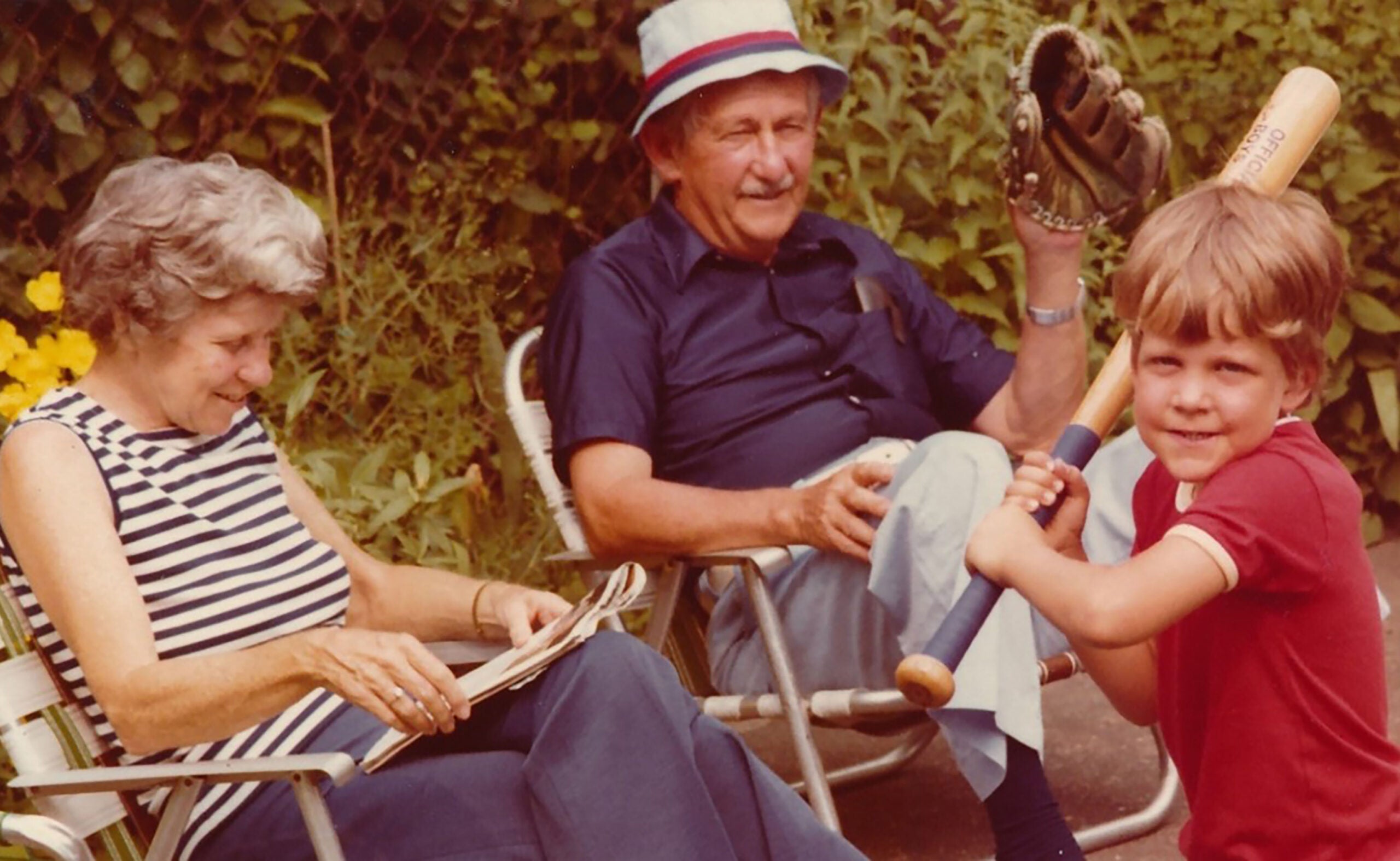 Smiling older man in a hat and dark shirt sits in a lawn chair holding a baseball glove, while a young boy in a red shirt poses with a bat, ready to swing.