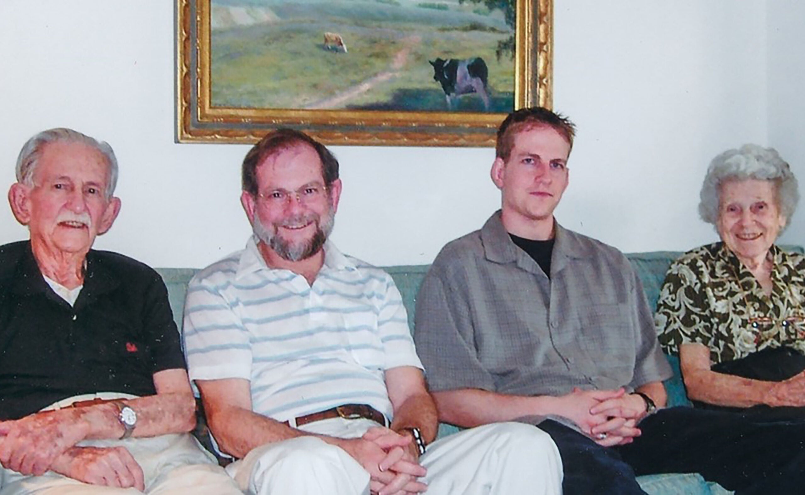 Four generations of a family sitting together on a couch, with two older adults on the ends and two younger men in the middle, all smiling at the camera.