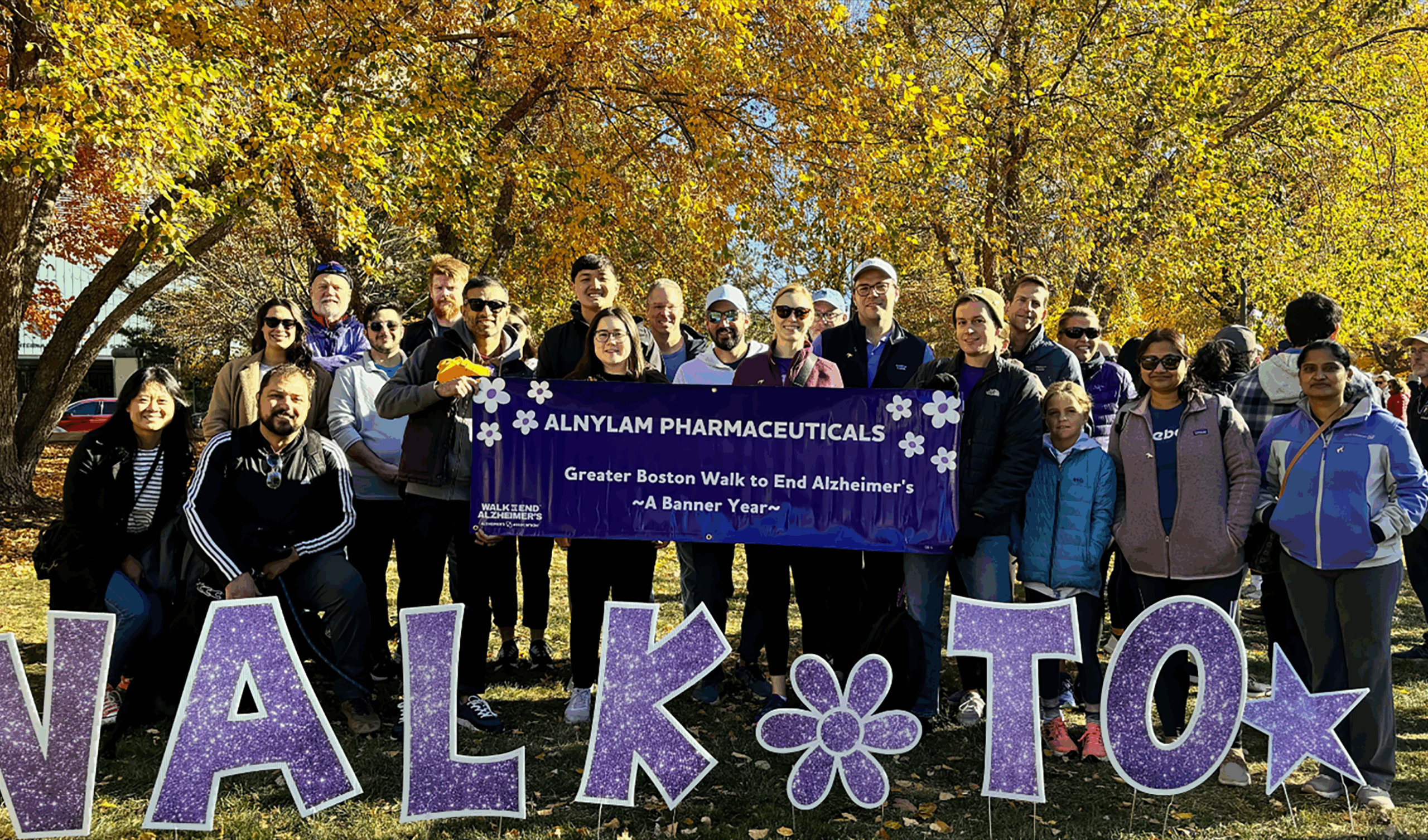A group of people from Alnylam Pharmaceuticals stand together outdoors holding a purple banner that reads “Greater Boston Walk to End Alzheimer’s A Banner Year” during the fall event, with colorful autumn trees in the background. Large purple glitter letters spelling “WALK TO” sit in front of the group.