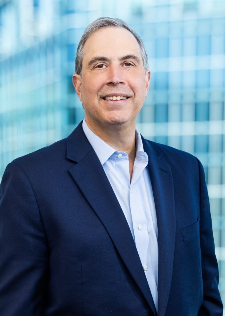 Professional headshot of a man in a navy blue blazer and light blue shirt, smiling in front of a blurred glass building background.