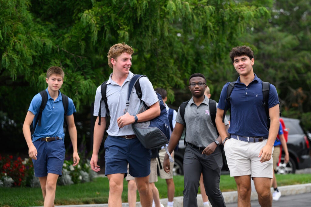 A group of high school boys wearing backpacks and collared shirts walk together outside, smiling and talking on a tree-lined path.