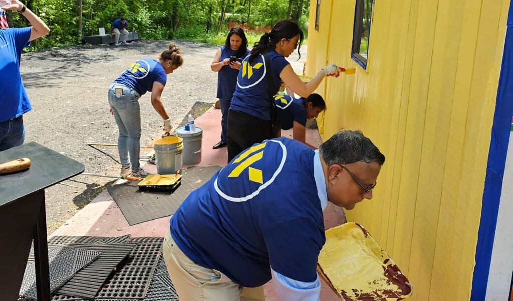 Group of Webster Bank employees in blue shirts with the company logo painting the exterior of a bright yellow building during a volunteer event.