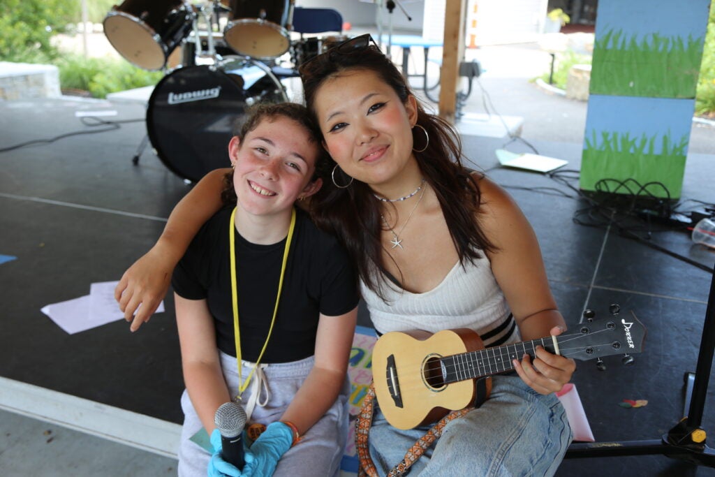 Two girls smiling, one holding a ukulele and the other one a microphone